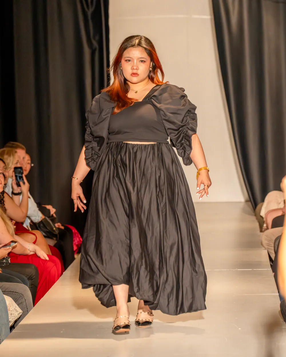 A model in a black outfit walking a runway between seated spectators