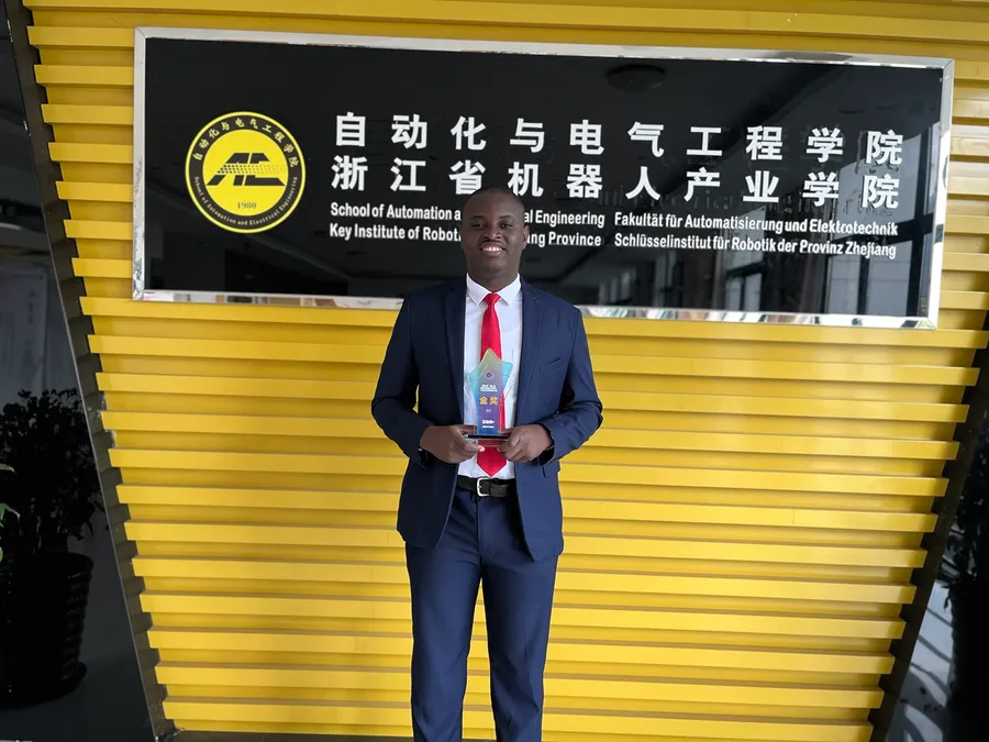 Kenneth in a suit holding an award in front of the sign of a Zhejiang automation and electrical engineering institute