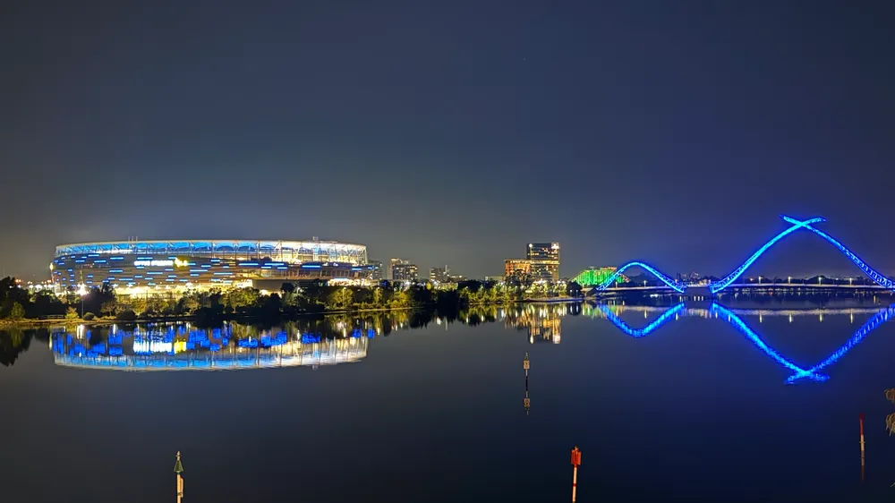 Optus Stadium and the Matagarup Bridge lit blue at night, reflected in the Swan River
