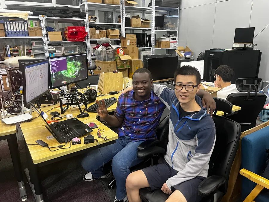 Kenneth and a colleague sitting at a desk in the Robsense lab, with monitors and drone parts on the shelves behind them