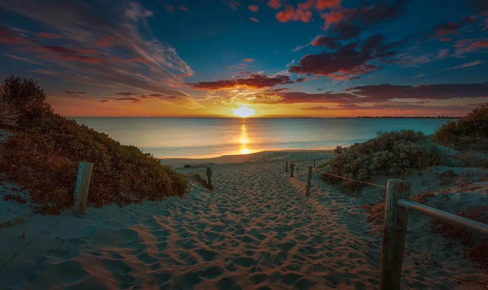 A sandy path between dunes leading to the sun setting over the ocean